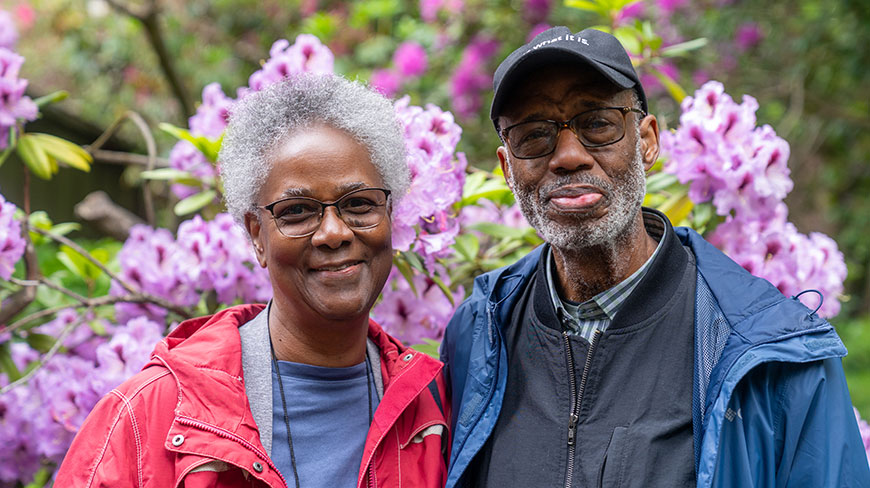 A smiling couple stands before blooming purple flowers in a rhododendron garden during an outdoor excursion in Utah.