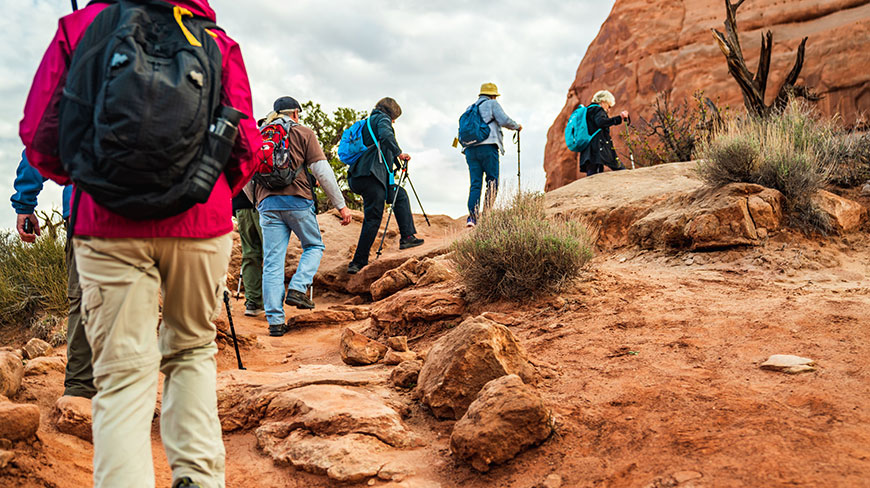 A group of hikers with backpacks and trekking poles ascend a rocky red trail during a Best of Utah hike.