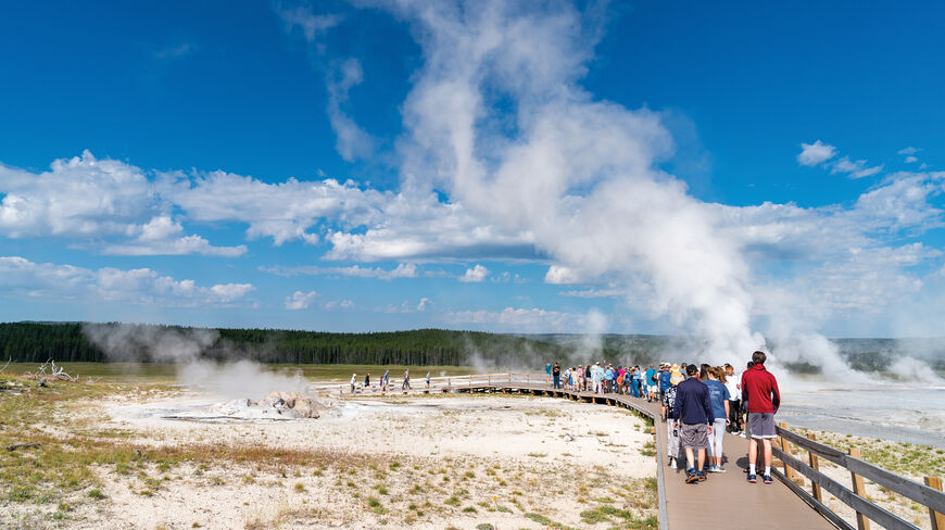 A group of travelers explore a steaming thermal basin from a boardwalk during an active adventure in Yellowstone National Park.
