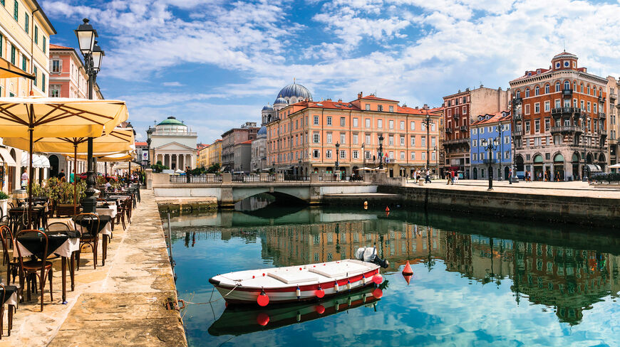 A white boat floats on the Grand Canal in Trieste, Italy, with colorful historic buildings and outdoor cafe seating reflected in the bright blue water.