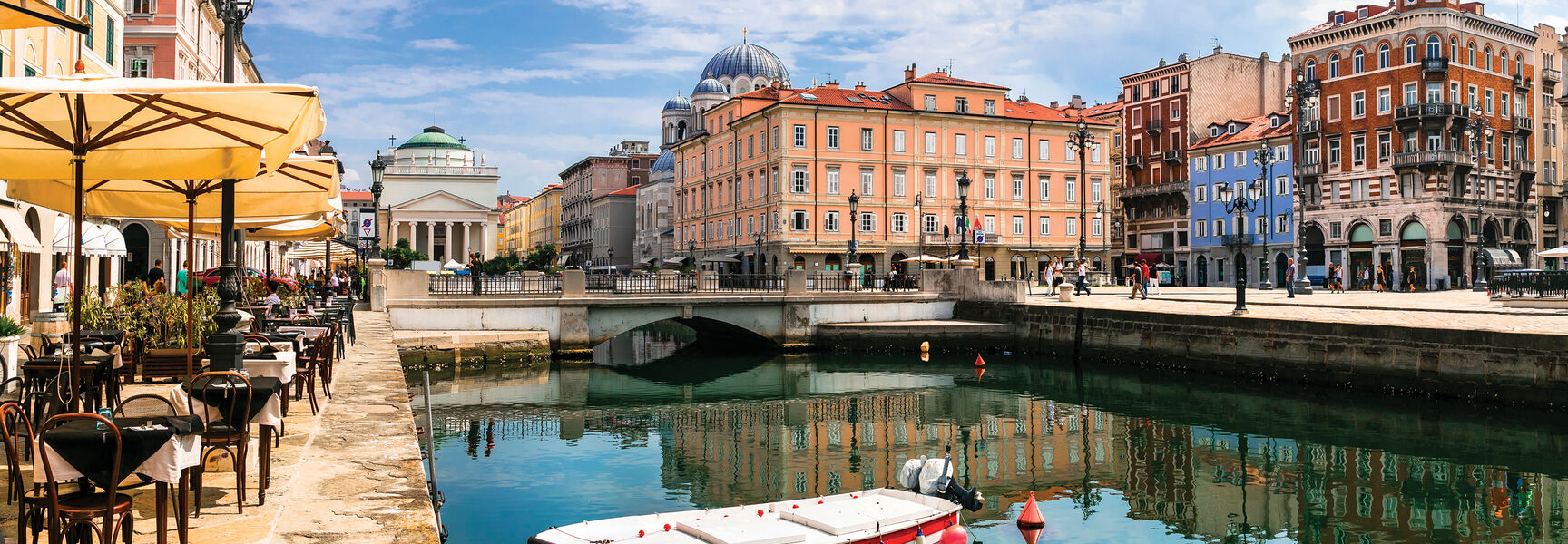 A white boat floats on the Grand Canal in Trieste, Italy, with colorful historic buildings and outdoor cafe seating reflected in the bright blue water.