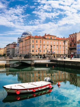 A white boat floats on the Grand Canal in Trieste, Italy, with colorful historic buildings and outdoor cafe seating reflected in the bright blue water.