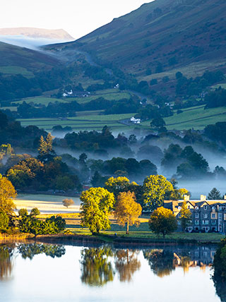 A misty morning in Grasmere, England, shows a still lake reflecting trees and a hotel in front of rolling green hills.