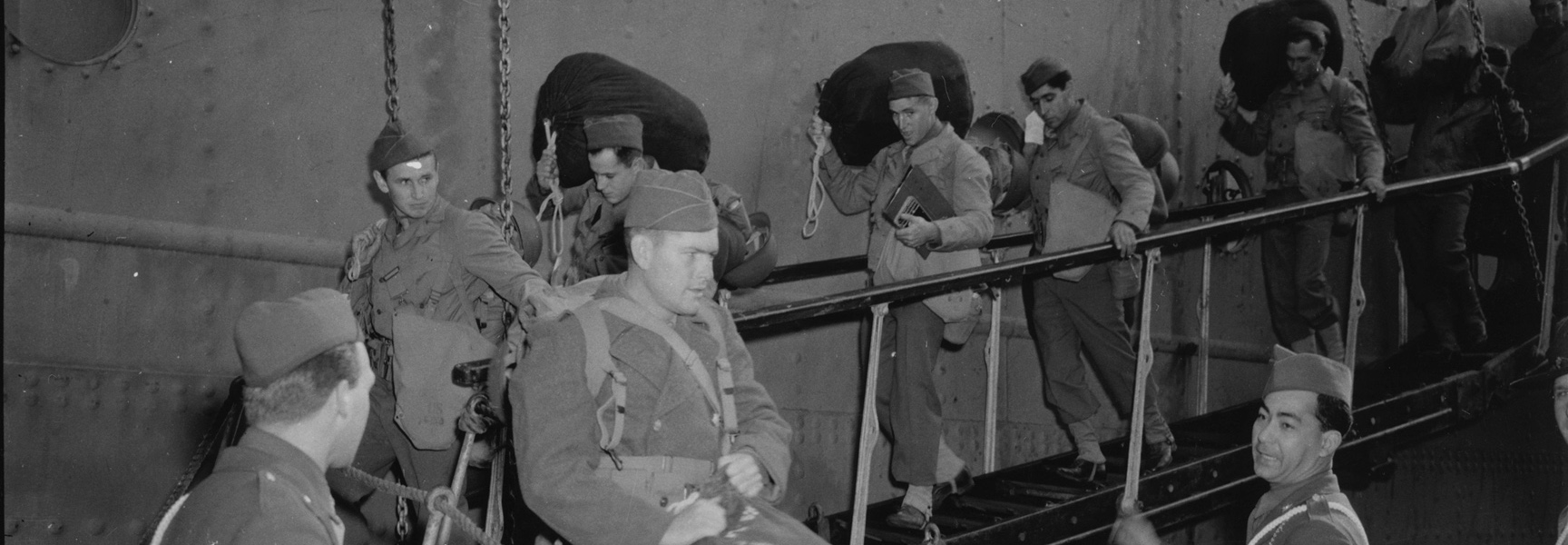 A black and white photograph shows American GIs with their bags disembarking a ship in Britain during World War II.