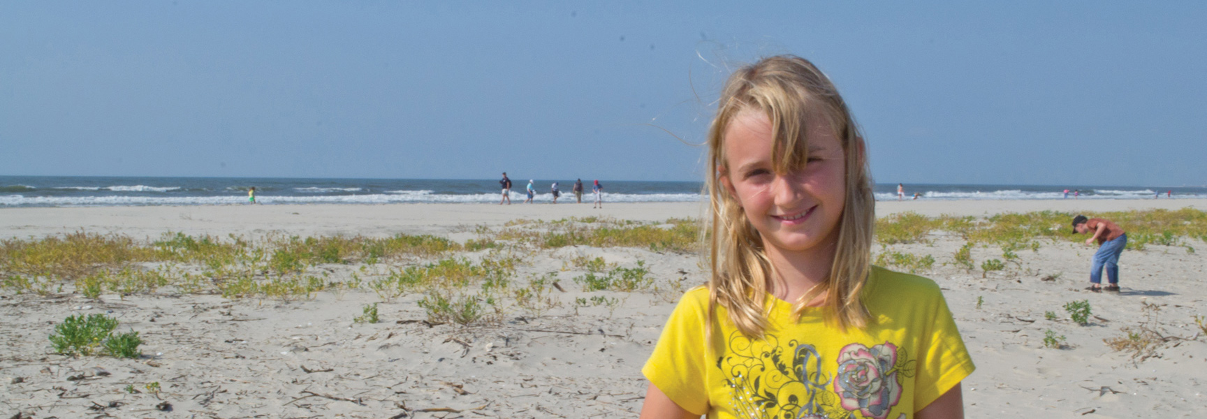 A young girl in a yellow shirt smiles on a sandy beach on one of Georgia's Barrier Islands, with the ocean behind her.