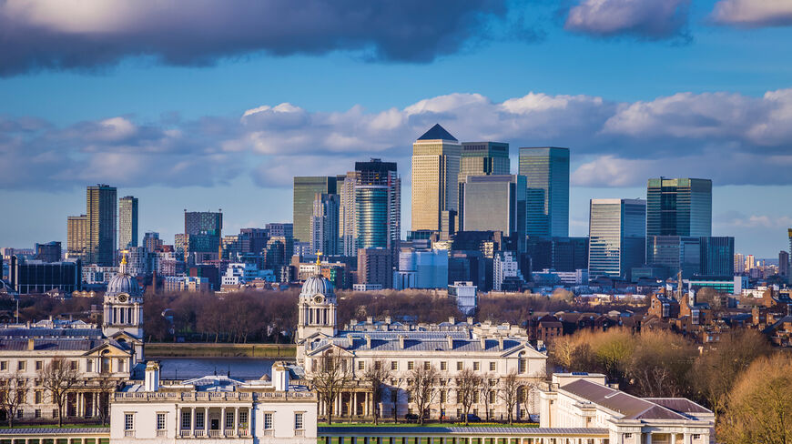 The historic Queen's House and Old Royal Naval College in London stand before the modern Canary Wharf skyline under a bright, cloudy sky.