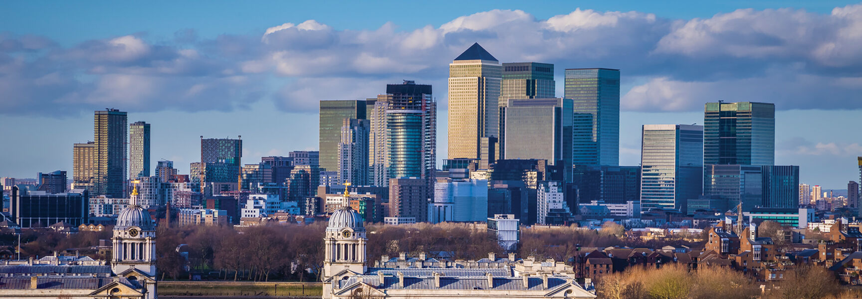 The historic Queen's House and Old Royal Naval College in London stand before the modern Canary Wharf skyline under a bright, cloudy sky.
