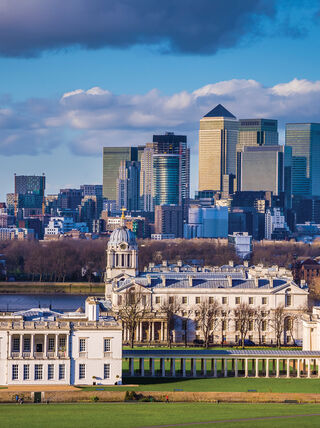 The historic Queen's House and Old Royal Naval College in London stand before the modern Canary Wharf skyline under a bright, cloudy sky.