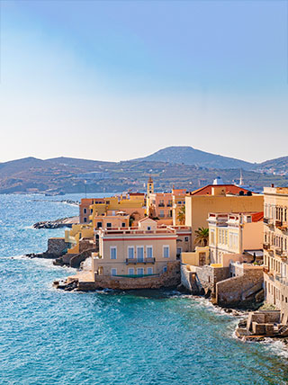 Colorful historic buildings line the rocky coast of the Aegean Sea in Ermoupoli, a charming port town on the Greek island of Syros.