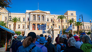 A group of travelers on a walking tour in Syros, Greece, gazes up at a large, neoclassical government building under a clear blue sky.