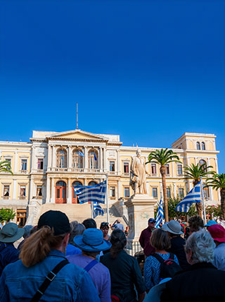 A crowd of tourists on a walking tour admire the grand City Hall building with Greek flags waving in Ermoupoli, Syros, Greece.