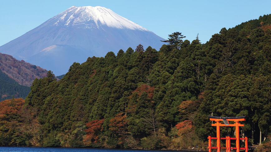 A red torii gate stands at the water's edge with snow-capped Mount Fuji rising behind forested hills along Japan's historic Tokaido Road.