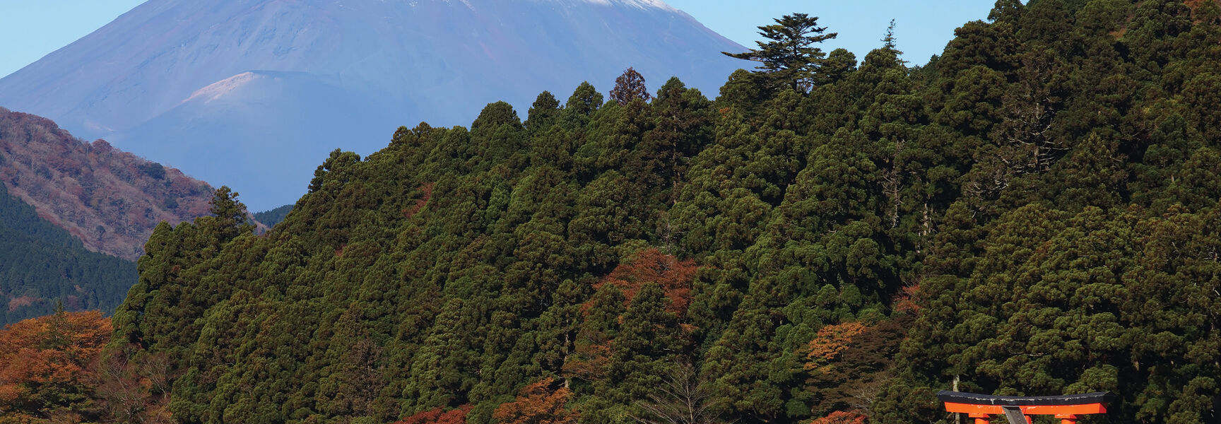 A red torii gate stands at the water's edge with snow-capped Mount Fuji rising behind forested hills along Japan's historic Tokaido Road.