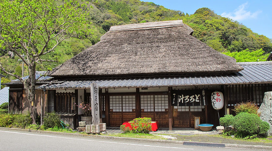 The historic Chojiya inn with a traditional thatched roof sits along the Tokaido Trail in Japan, nestled against a lush, green hillside.