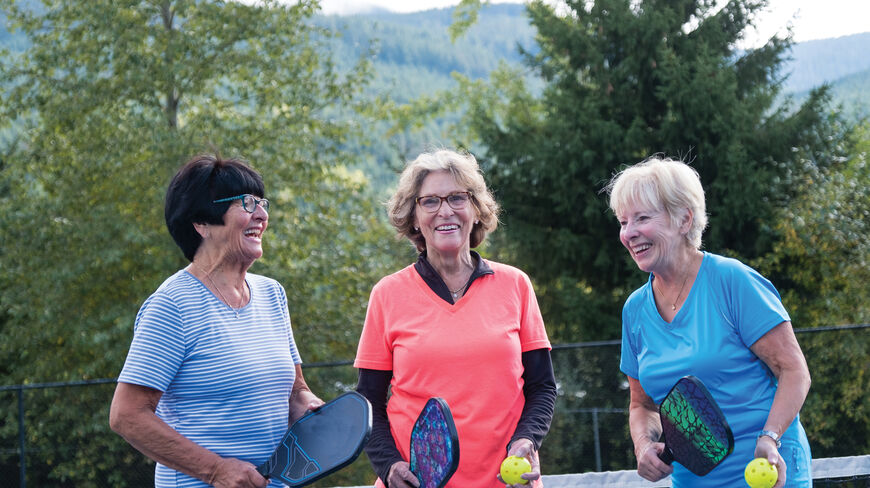 A pickleball group of three senior women laughing while holding paddles and balls on an outdoor court in Washington.