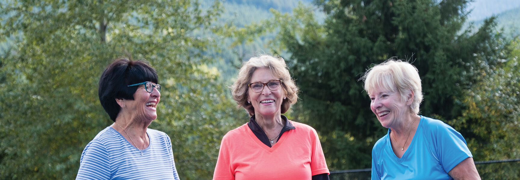 A pickleball group of three senior women laughing while holding paddles and balls on an outdoor court in Washington.