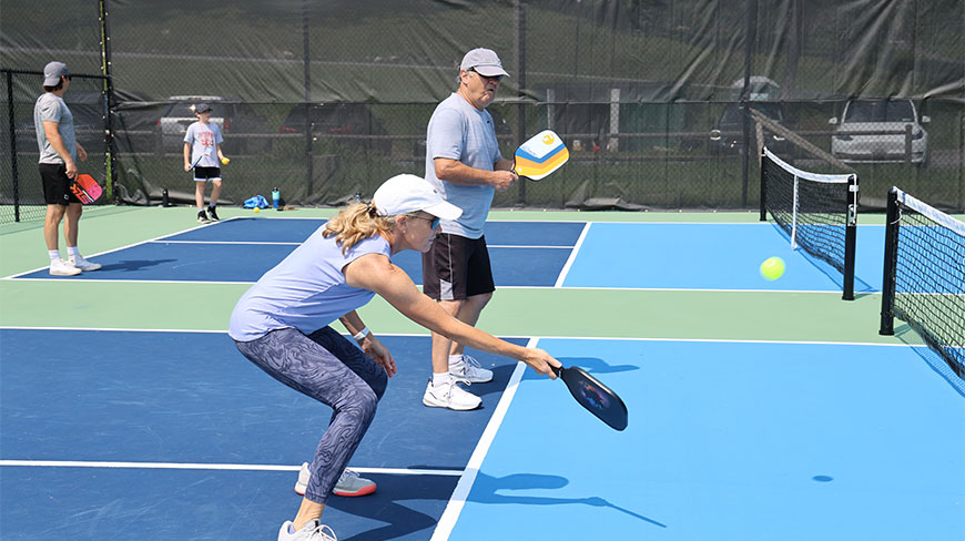 Pickleball players compete on an outdoor blue and green court in New York, USA, with a woman reaching to hit the ball.