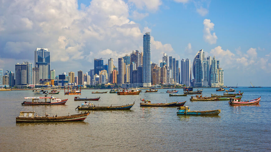 The Panama City skyline features modern skyscrapers rising above a bay filled with small fishing boats under a cloudy blue sky.