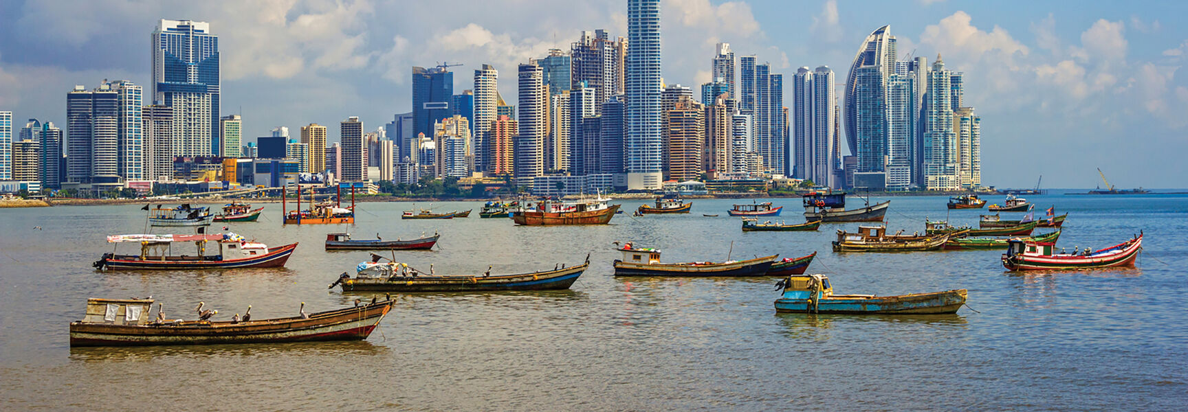 The Panama City skyline features modern skyscrapers rising above a bay filled with small fishing boats under a cloudy blue sky.