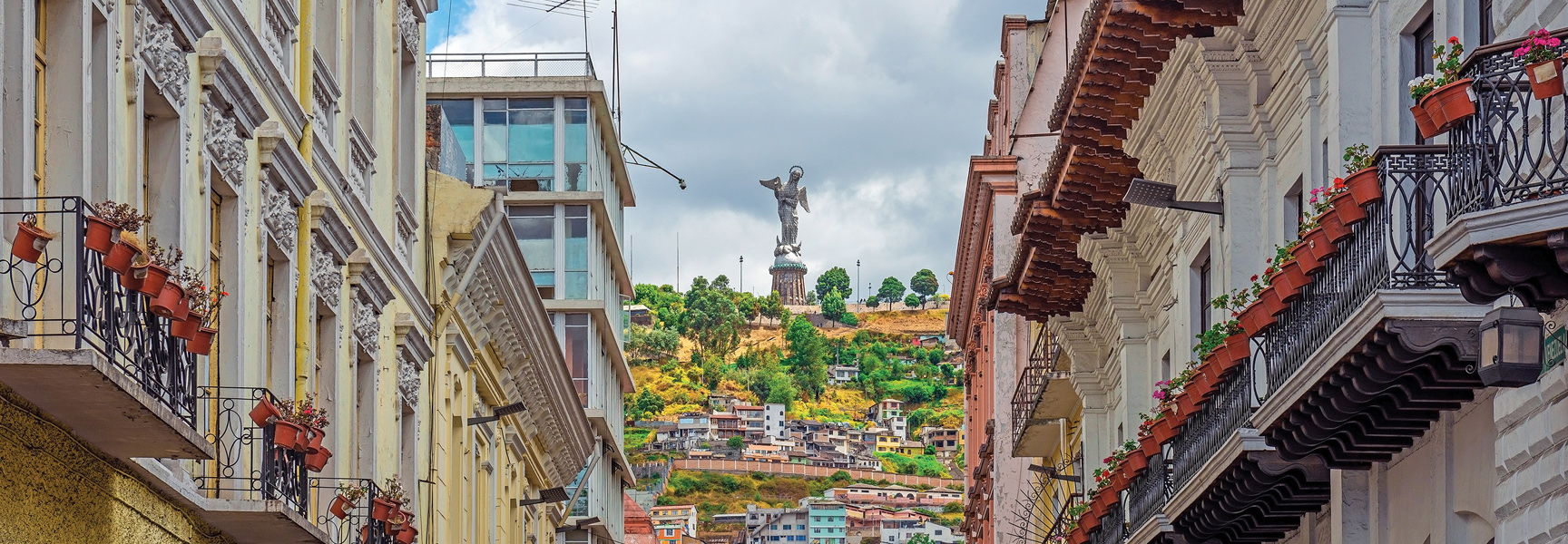 A view down a street in Quito, Ecuador, reveals the Virgin of El Panecillo statue standing on a green hill between historic apartment buildings.