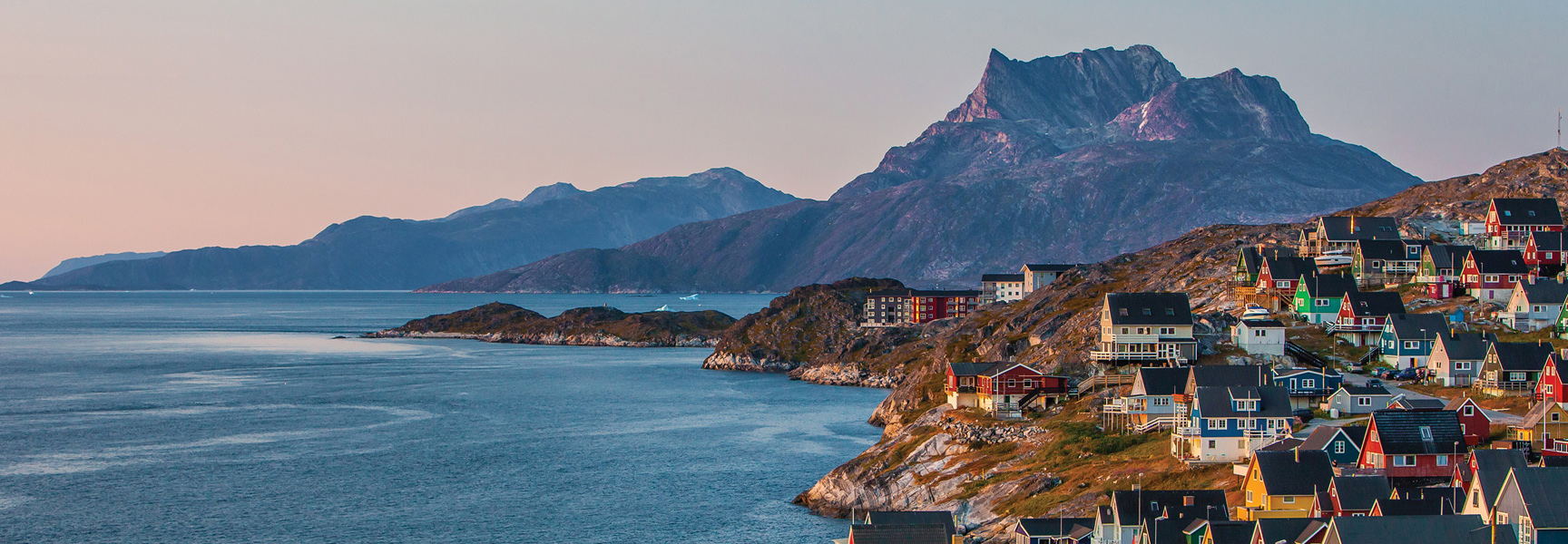 Colorful houses are scattered across a rocky hillside along the coast of Greenland, with large mountains in the background under a soft-lit sky.