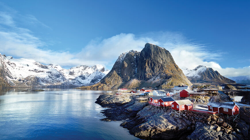 Red houses on stilts line the rocky shore of a Norwegian fjord beneath towering snow-capped mountains under a bright blue Arctic sky.