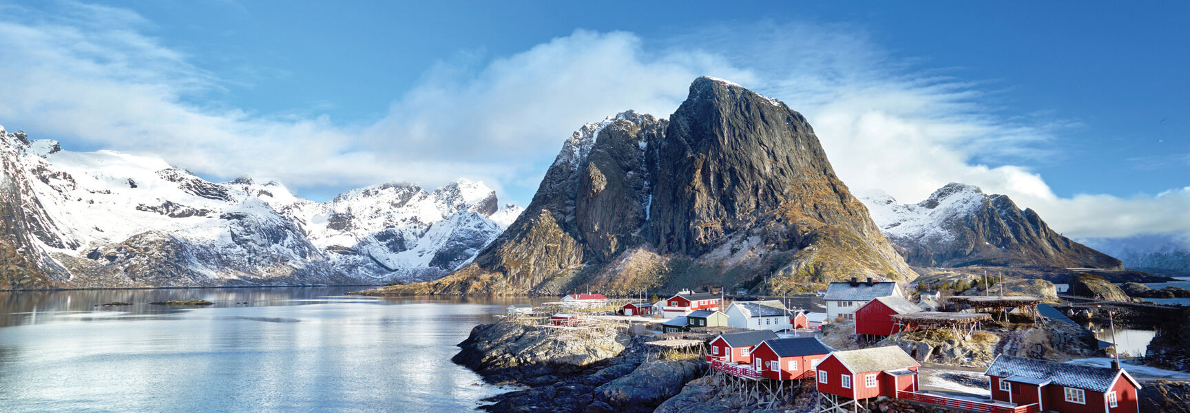 Red houses on stilts line the rocky shore of a Norwegian fjord beneath towering snow-capped mountains under a bright blue Arctic sky.