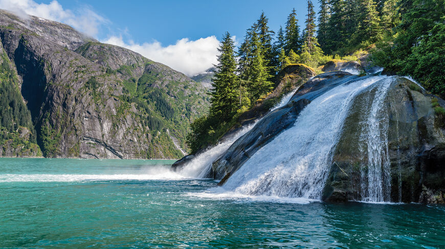 A scenic waterfall cascades over rocks into turquoise water along Alaska’s Inside Passage, surrounded by steep mountains and the Tongass National Forest.