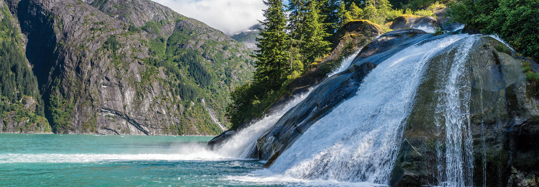 A scenic waterfall cascades over rocks into turquoise water along Alaska’s Inside Passage, surrounded by steep mountains and the Tongass National Forest.
