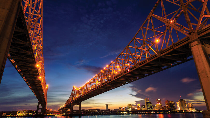 The illuminated bridge spans the Mississippi River at dusk with the New Orleans skyline glowing in the background.
