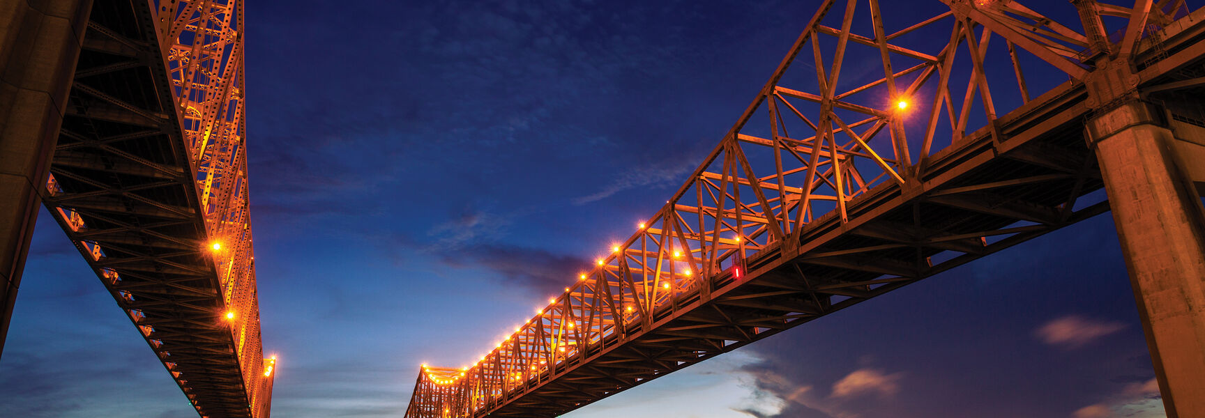 The illuminated bridge spans the Mississippi River at dusk with the New Orleans skyline glowing in the background.