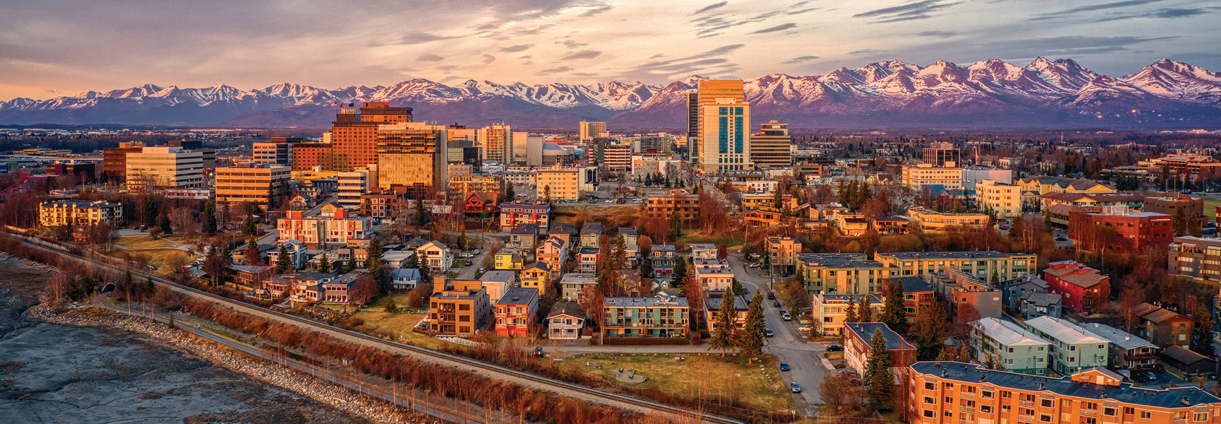 An aerial view of the Anchorage, Alaska skyline at sunset, with the snow-covered Chugach Mountains in the distance.