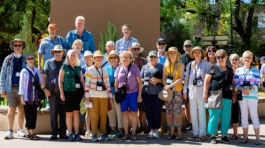 A large group of smiling older adults wearing casual clothing and hats pose for a photo outdoors on a sunny day.