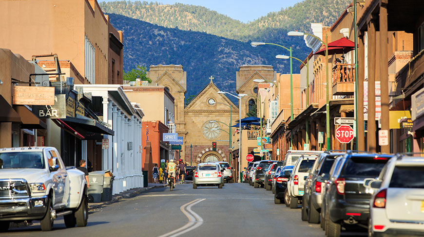 A street lined with adobe buildings in Santa Fe, New Mexico, leads towards a historic cathedral with mountains visible in the distance.