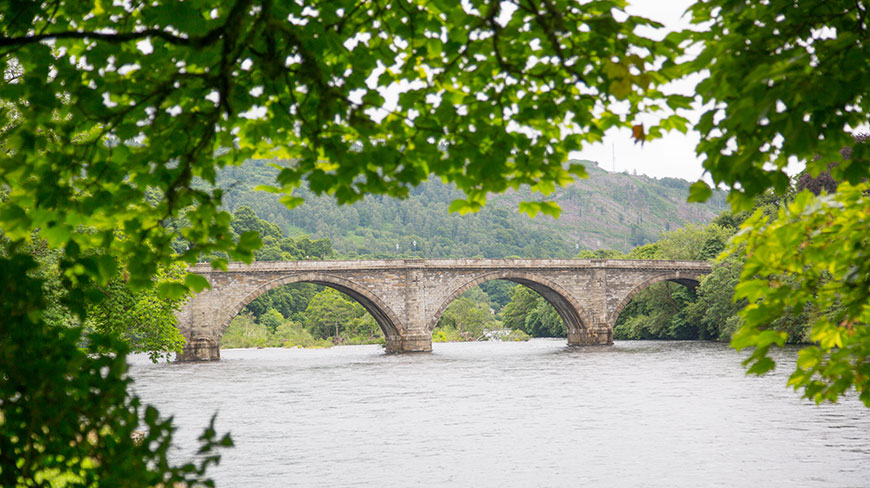 The historic multi-arched stone Dunkeld Bridge in Scotland, framed by lush green leaves, spans a wide river with forested hills in the background.