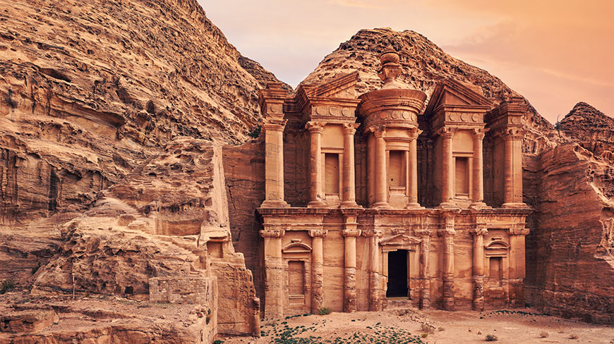 The ornate facade of the Ad Deir Monastery is carved into a sandstone mountain in Petra, Jordan, under a warm-hued sky.