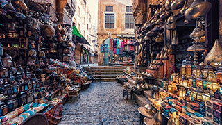 A bustling, narrow cobblestone street in a Cairo market is lined with shops selling traditional lanterns, souvenirs, and other beautiful wares.
