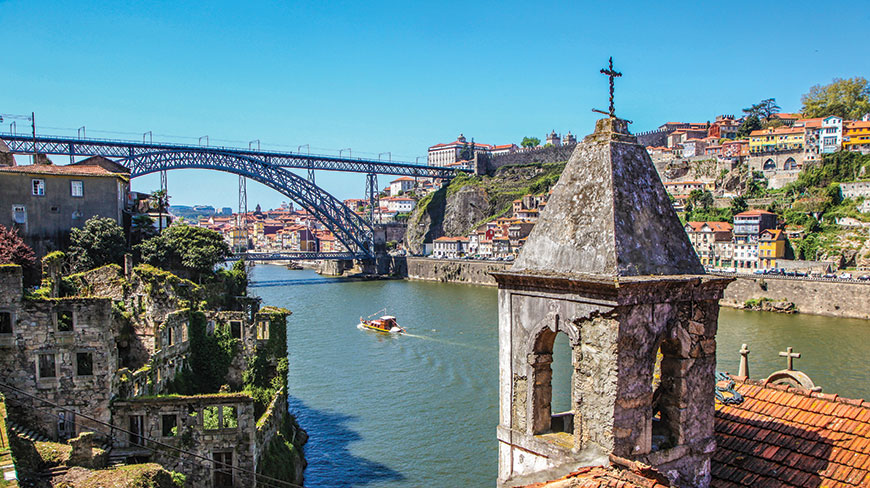 A stone bell tower overlooks the scenic Douro River and the Dom Luís I Bridge in the city of Porto on a sunny day.