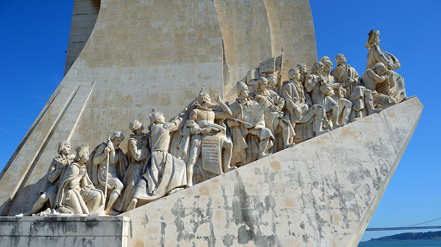The stone-carved prow of the Monument to the Discoveries in Lisbon, crowded with historical figures, juts out under a brilliant blue sky.