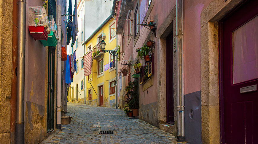 A narrow cobblestone alleyway slopes up between colorful buildings in Lisbon, with laundry and birdcages hanging from the windows.