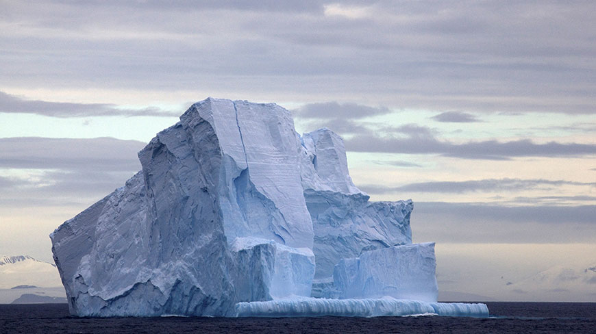 A large, craggy iceberg floats on the dark ocean water under a cloudy, overcast sky.