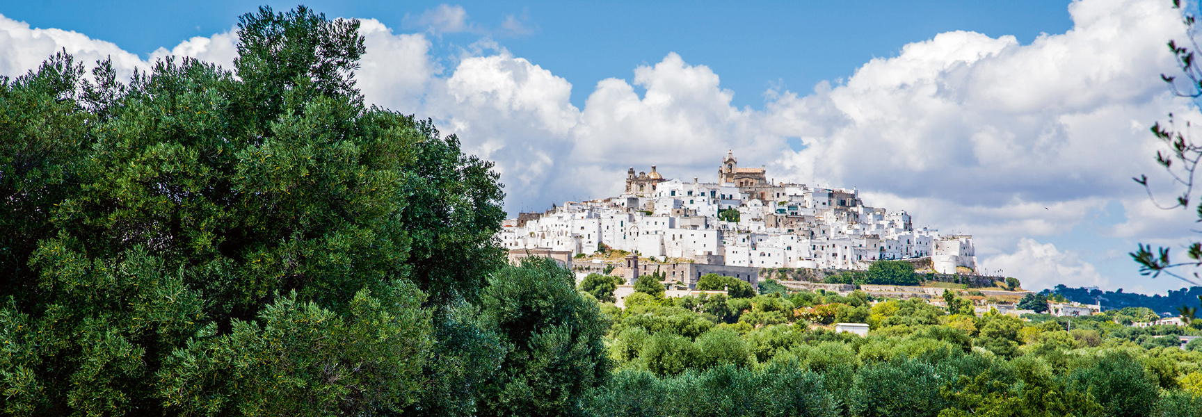 A whitewashed town on a hill in Puglia, Italy, is surrounded by lush green trees under a blue sky with white clouds.