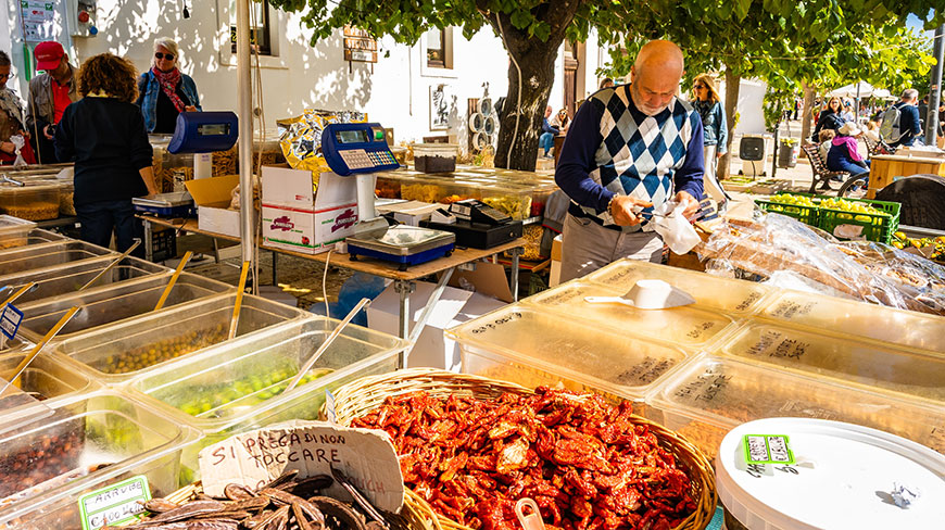 A vendor serves customers at a sunny outdoor market stall filled with bins of olives, nuts, and sun-dried tomatoes under a leafy tree.