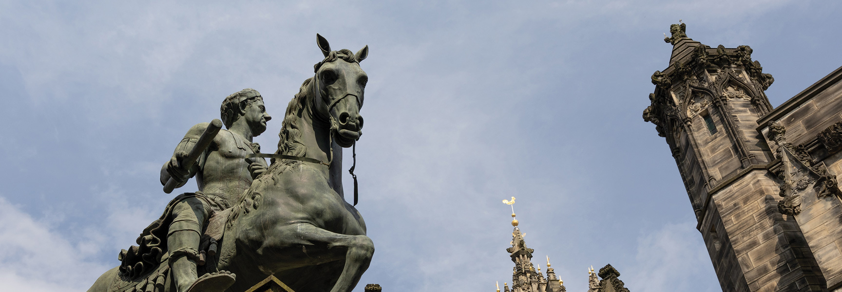 A low-angle view of an equestrian statue next to a historic stone building against a partly cloudy sky.