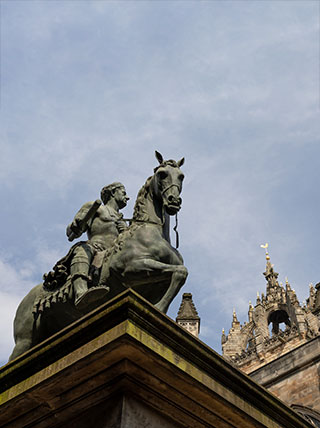 A low-angle shot of a bronze equestrian statue on a stone pedestal with the top of a historic building in the background.