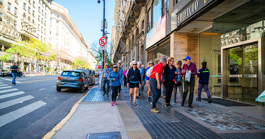 A group of travelers on a walking tour follow their guide along a sunny city sidewalk in Buenos Aires.