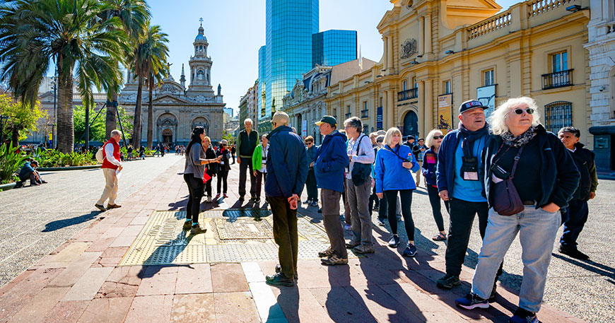 A group of tourists stands on a sunny plaza in Santiago, Chile, listening to a tour guide with historic buildings and palm trees behind them.