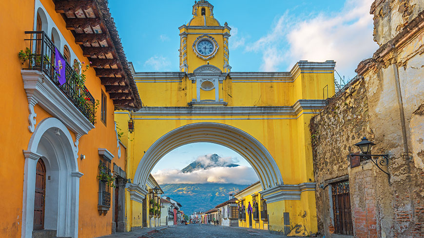 The yellow Santa Catalina Archway in Antigua frames a view of a volcano rising in the distance above a cobblestone street.