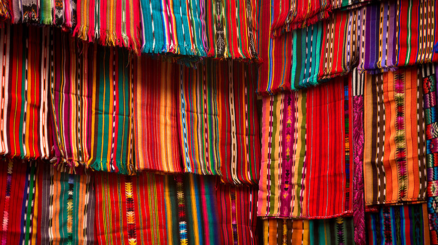 A vibrant, layered display of colorful, hand-woven striped textiles with fringed edges.