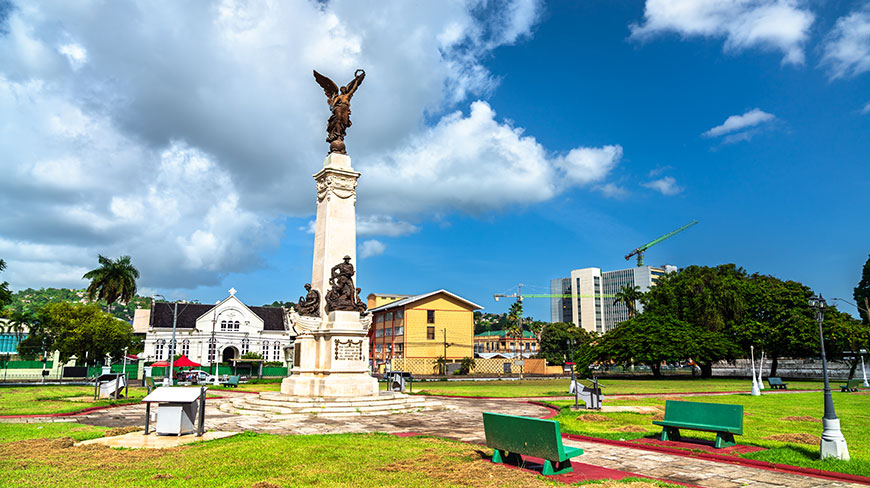 A tall, winged statue monument stands in the grassy Memorial Park in Port of Spain under a partly cloudy sky with city buildings behind.